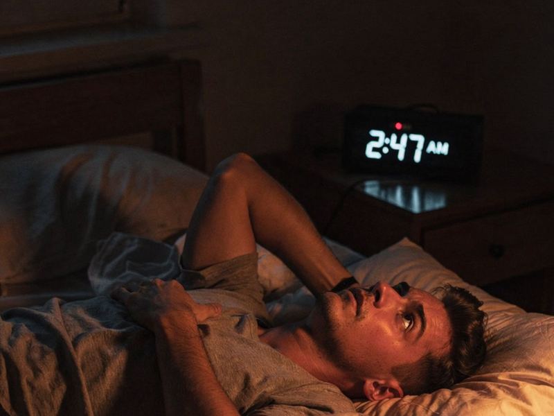 A man awake in bed next to a clock showing 2:47 AM